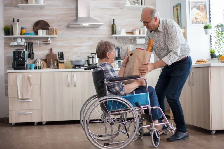 senior-man-helping-his-wife-by-taking-grocery-paper-bag-from-her-mature-people-with-fresh-vegetables-from-market-living-with-disabled-person-with-walking-disabilities-min-scaled